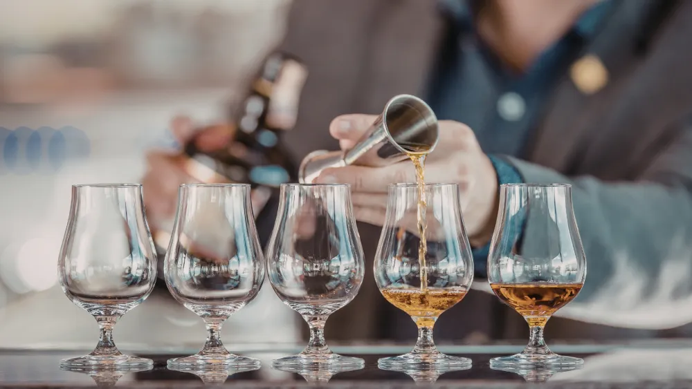 A bartender pours whiskey into a row of tulip glasses on a bar counter, with a line of amber liquid forming as he tilts the bottle.