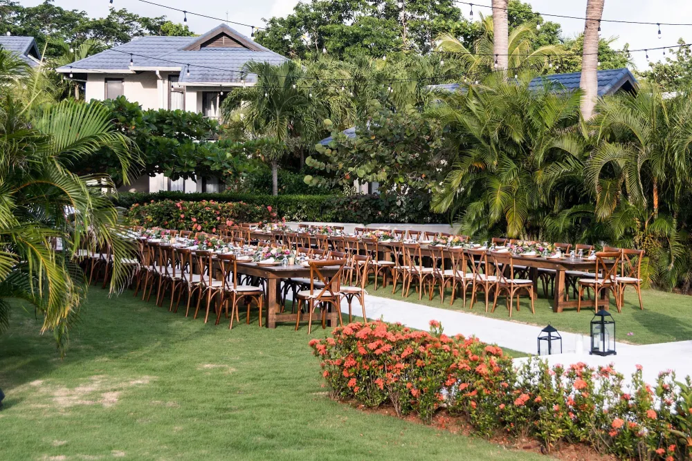 A long row of wooden benches is arranged under a roofed shelter beside a grassy yard, with trees and a white building in the background.