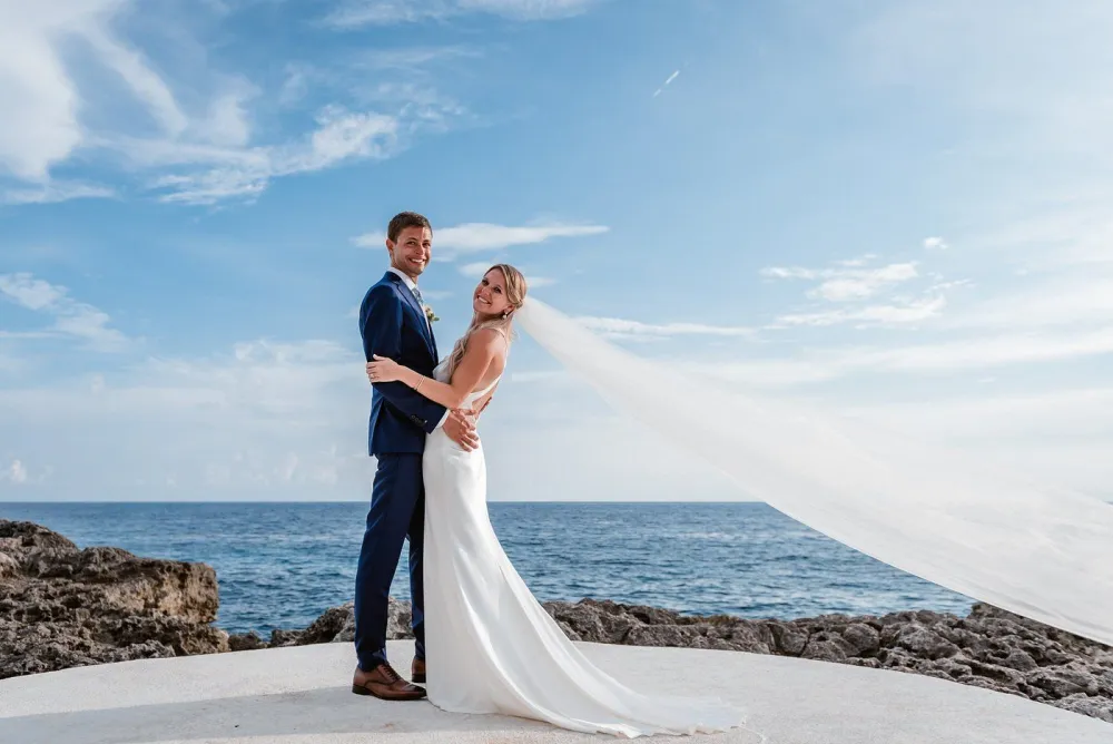 A bride and groom kiss on the beach, holding each other in wedding attire with a sunny, blue sky and calm sea behind them.