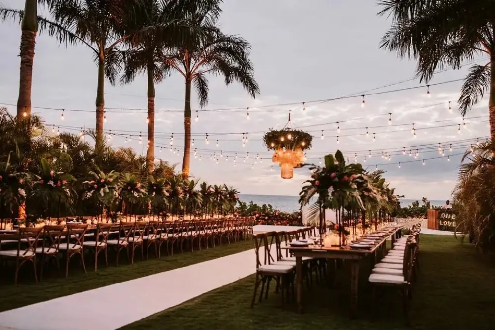 Outdoor wedding reception setup with long tables, string lights, palm trees, and a dramatic hanging flame lantern adorning the aisle, creating a warm festive vibe.