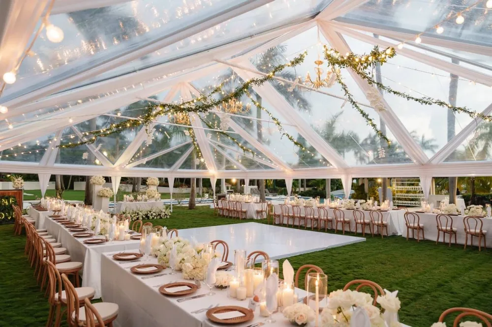 A modern outdoor wedding venue with white chairs, wooden arbors, string lights, and greenery under a clear glass canopy.