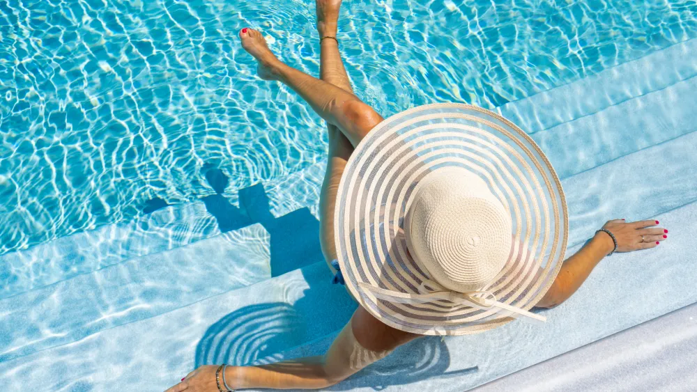 A person in a large striped sunhat sits at the pool edge, with legs in the water and bright blue pool surroundings.