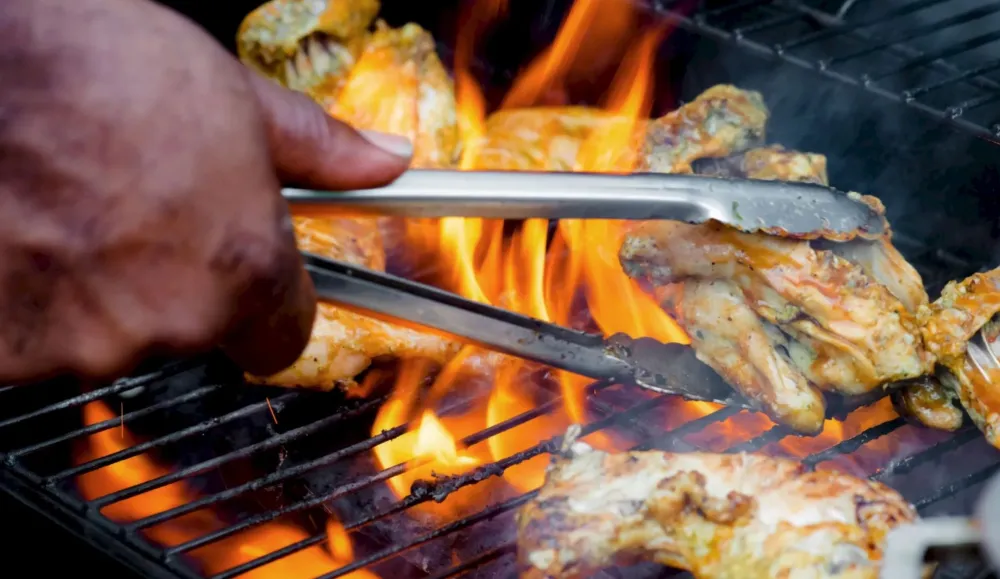 Grilled chicken pieces sizzle over open flames on a charcoal grill, held near the fire by a hand and tongs.