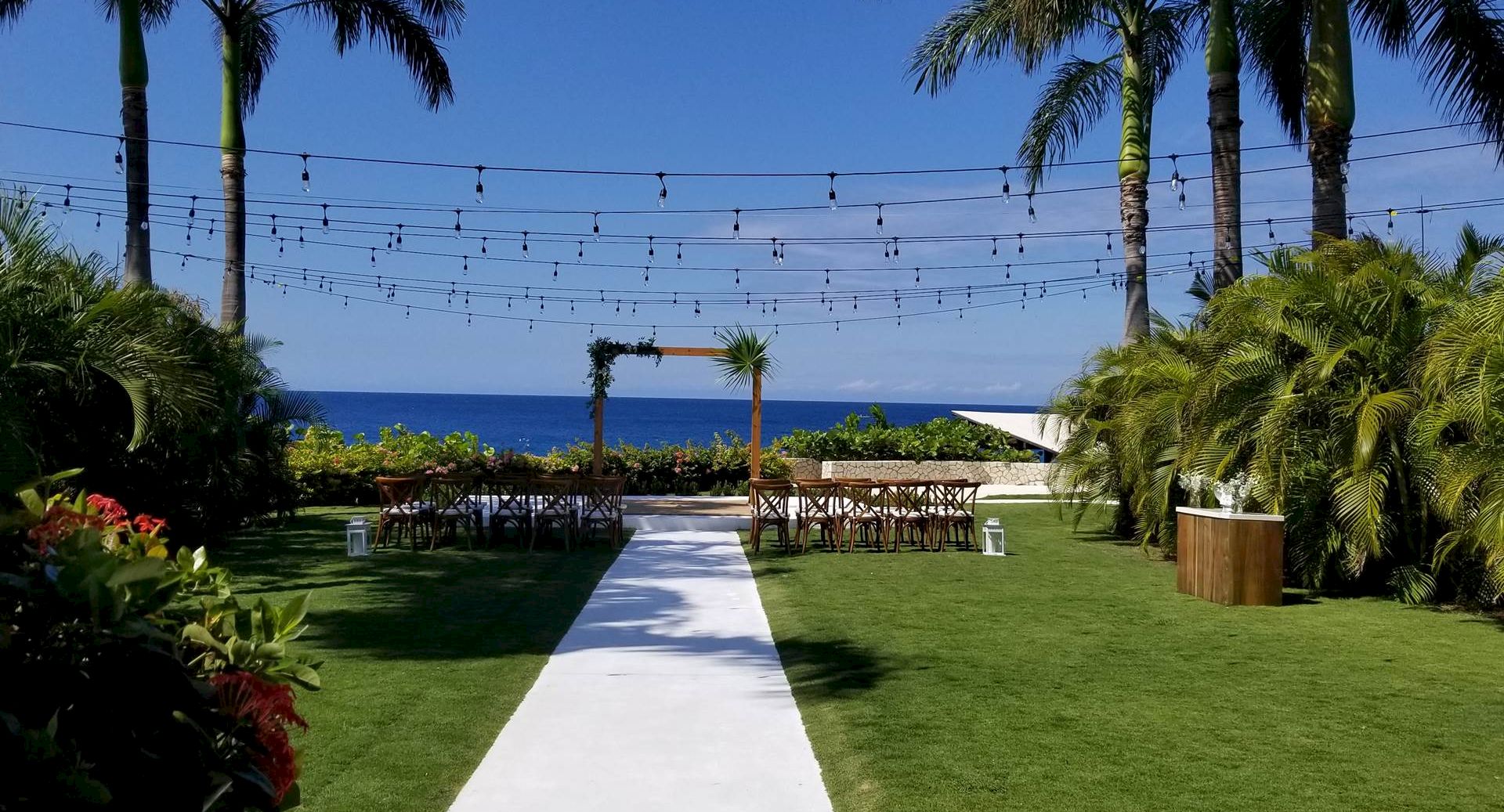 A beachside wedding setup with string lights, a white aisle leading to an arch, surrounded by palm trees and lush greenery by the ocean.