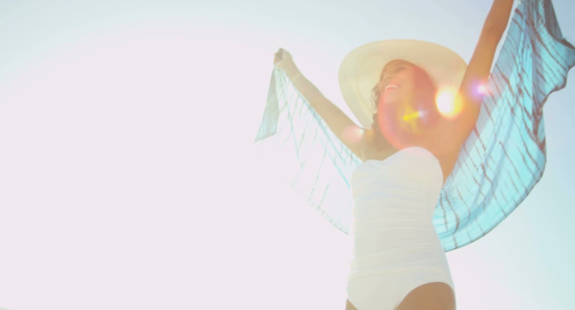 A woman in a white swimsuit stands under bright sun, wearing a wide-brim hat and a sheer blue cover, arms raised joyfully.