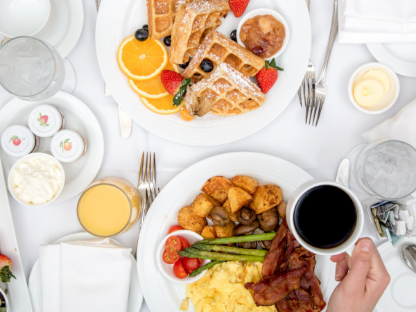 A breakfast spread with waffles, croissants, eggs, bacon, pancakes, fruit, pastries, coffee, orange juice, and a colorful plate of sides.
