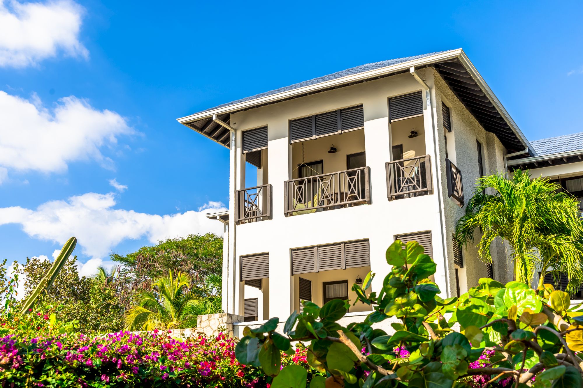 A two-story house with beige walls, dark shutters, and a small balcony, surrounded by lush tropical plants and bright blue sky.
