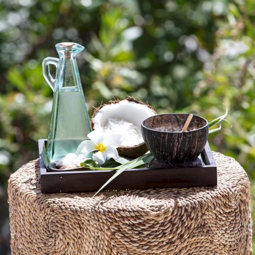 A glass bottle with a narrow neck on a wooden tray, plus a small wooden bowl and a coconut husk, placed on a woven stool outdoors, with greenery in the background.