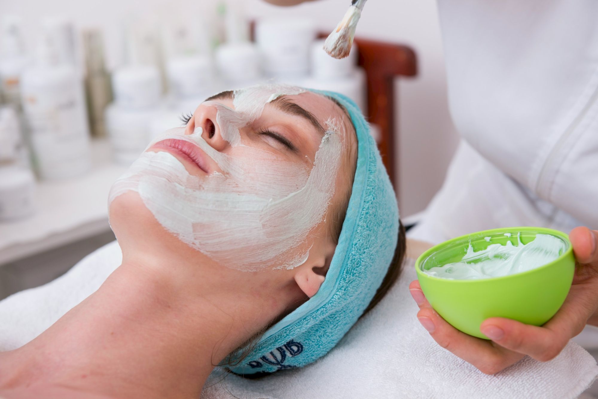 A woman lies with a facial mask applied, eyes closed, while a therapist holds a green bowl of cream near her face.