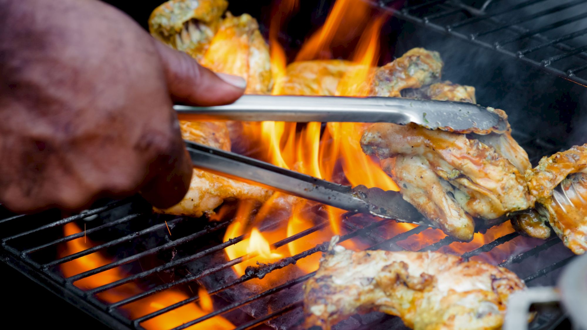 A cook uses tongs to place grilled chicken over an open flame on a barbecue grill.