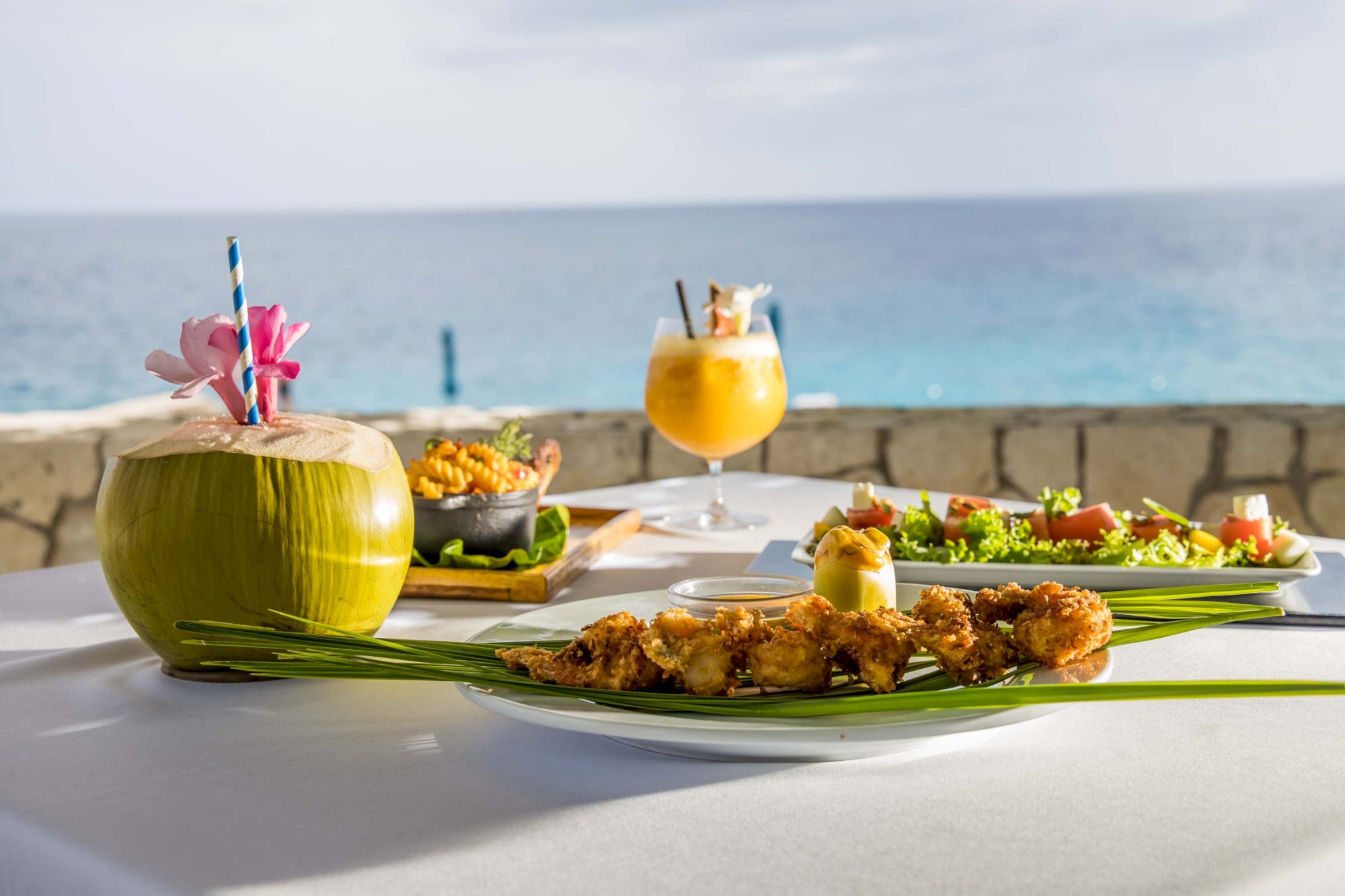 A tropical meal by the sea: a coconut drink, colorful cocktails, and a variety of fresh seafood plates on a seaside table.