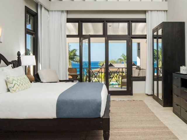 A modern hotel room with a dark wood bed, white and gray linens, a glass door balcony overlooks palm trees and the ocean, bright and airy.