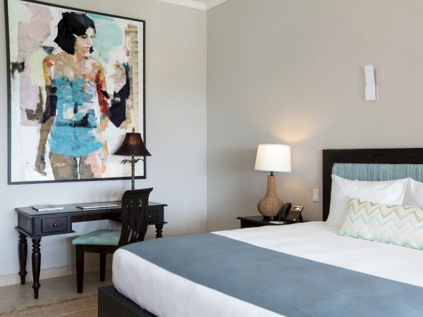 A modern hotel room with a queen bed, white linens, blue throw, two lamps, a framed artwork, and a small desk with a chair in the corner.