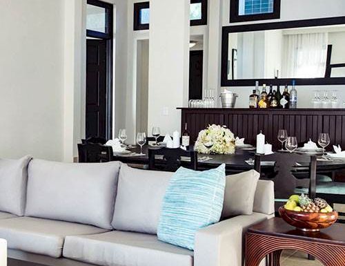 A modern, open living area with a light gray sofa, blue accent pillow, and a stylish dining space in the background. The room looks bright and elegant.