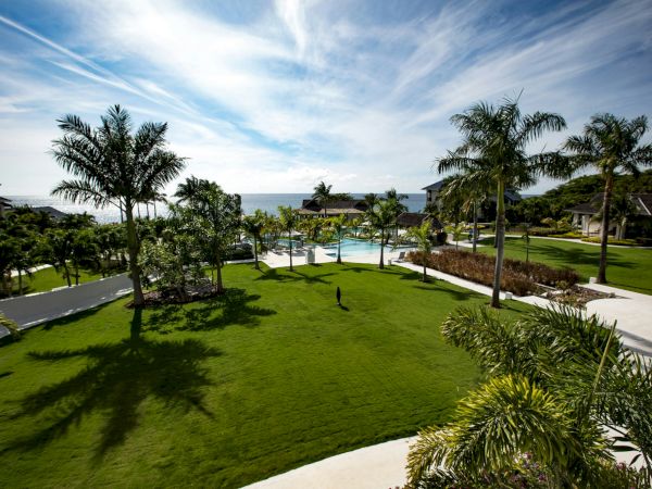 A sunny resort lawn with palm trees, a winding path, and a pool area near the coastline; bright sky, blue water, and manicured greenery.