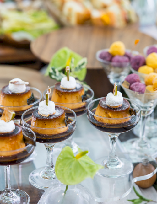 A fancy dessert spread: caramel-topped treats on glass cups, mini pies with dollops of cream, assorted fruit and nuts, and elegant flowers. End.