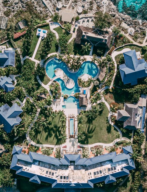 An aerial view of a tropical resort with blue pools, sun loungers, and lush gardens surrounding villas and cabanas.