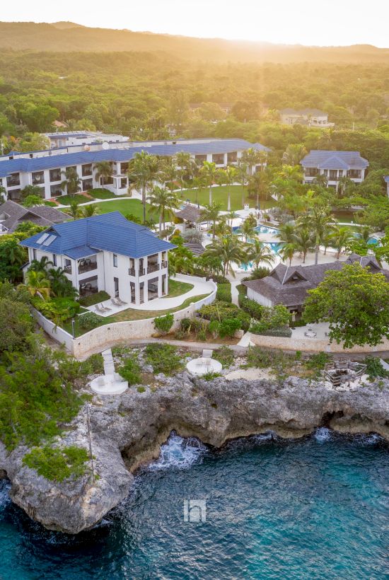 Aerial view of a tropical beachfront resort with blue-roof villas, lush palms, and a rocky cove at sunset.