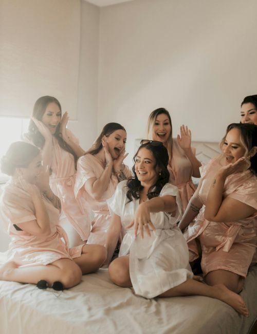 A group of bridesmaids gather on a bed, smiling and posing playfully for a photo, wearing matching light pink robes and one in white, in a cozy room.