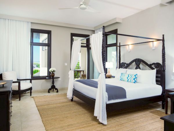 A luxurious tropical hotel room with a dark four-poster bed, white curtains, blue and white bedding, a rug, desk, chairs, and large windows with sheer curtains.