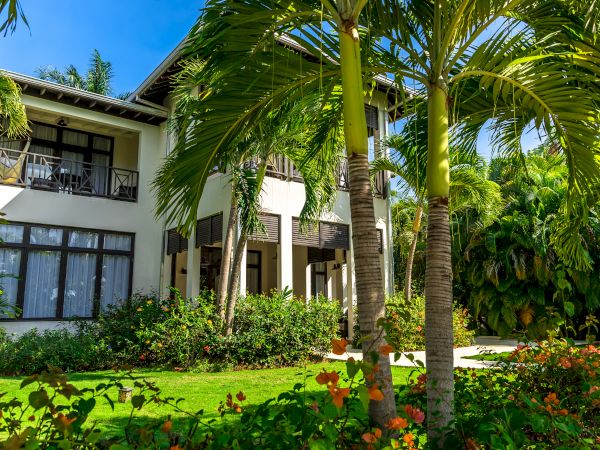 A tropical resort with a two-story white villa, lush green lawn, palm trees, colorful flowers, and sunny blue skies.