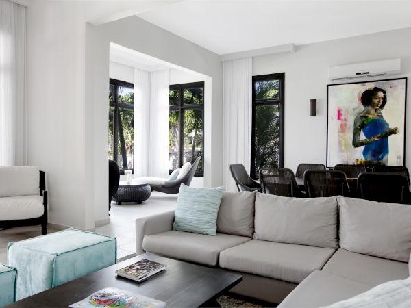 A bright modern living room with a gray sectional sofa, teal accents, large windows, and a colorful painting on the wall, overlooking a sunny seating area.