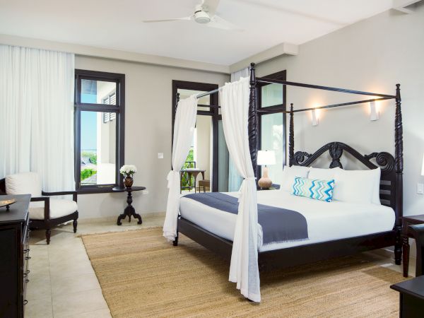 A luxurious bedroom with a four-poster bed, white canopy curtains, neutral tones, a beige rug, two nightstands, a desk and chair, and large windows letting in natural light.