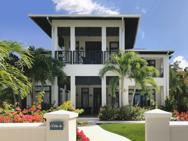 A modern two-story white villa with dark trim, lush tropical landscaping, palm trees, and a curved walkway leading to the entrance.