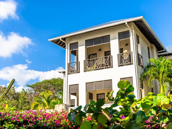 A two-story house with balconies, surrounded by lush tropical plants and a blue sky.