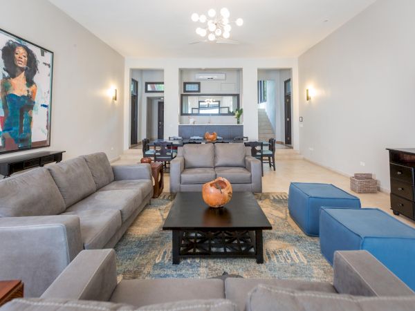 A modern, airy living room with gray couches, a black coffee table, blue ottomans, wall art, a TV, and a bright, open dining/kitchen area.