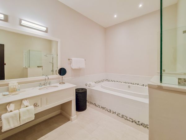 A modern bathroom with a double sink vanity, large mirror, wall lights, a bathtub with decorative tile border, towels, and a glass shower; clean and airy.