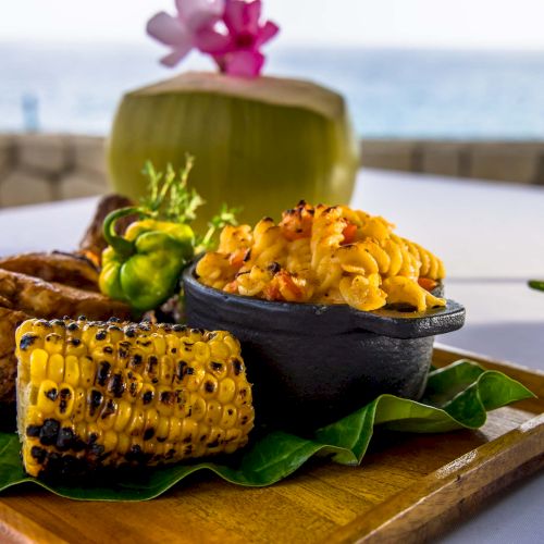 A tropical seafood dish with grilled corn, roasted plantain, and a saffron-tleck rice or side, served on a wooden board with a coconut drink, by the sea.