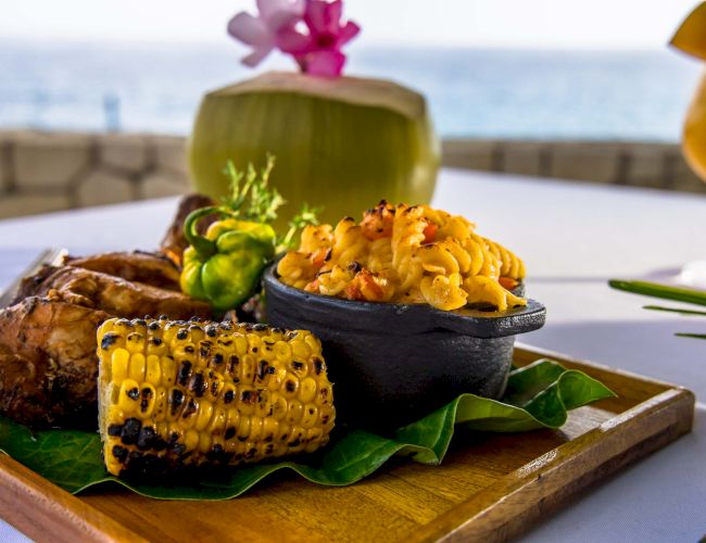 A tropical seafood dish with grilled corn, roasted plantain, and a saffron-tleck rice or side, served on a wooden board with a coconut drink, by the sea.