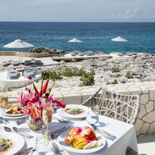 A seaside restaurant table set with white tablecloths, plates of food, flowers, and a view of the rocky shore and blue sea under a sunny sky.