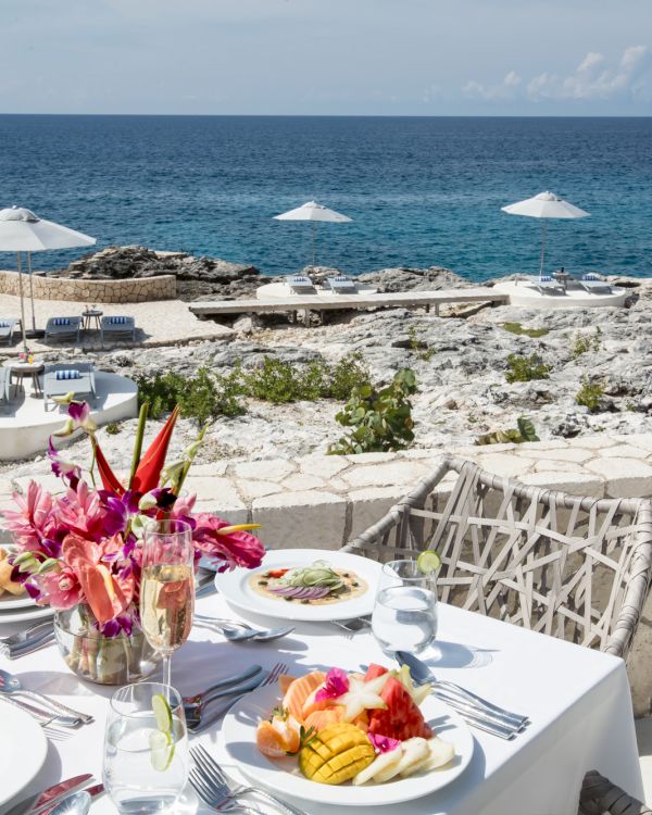 A seaside restaurant table set with white tablecloths, plates of food, flowers, and a view of the rocky shore and blue sea under a sunny sky.