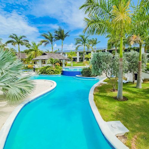 A tropical resort pool winds through palm trees toward a bright blue sky, with a curved slide and turquoise water, inviting relaxation.