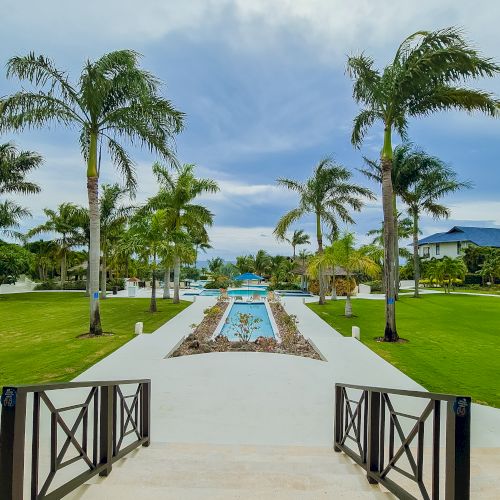 A tropical resort scene with a stone path leading to a pool, flanked by palm trees under a sunny sky, inviting relaxation and sunshine.