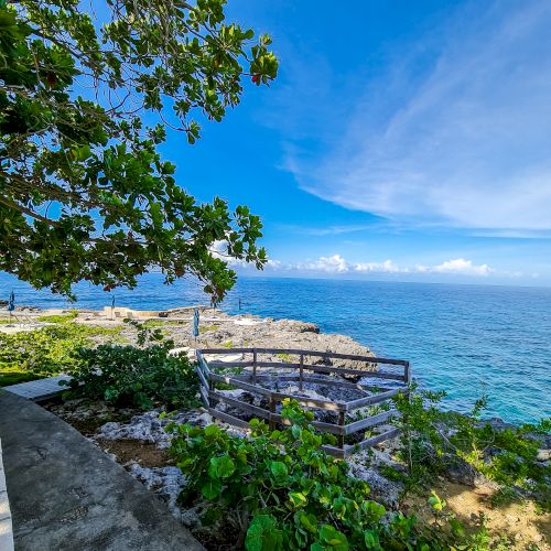A seaside scene with a stone wall, lush greenery, and a bright blue sky over calm water, creating a tranquil coastal view.