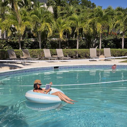 Two people on an inflatable raft in a bright pool, surrounded by palm trees and a sunny resort scene.