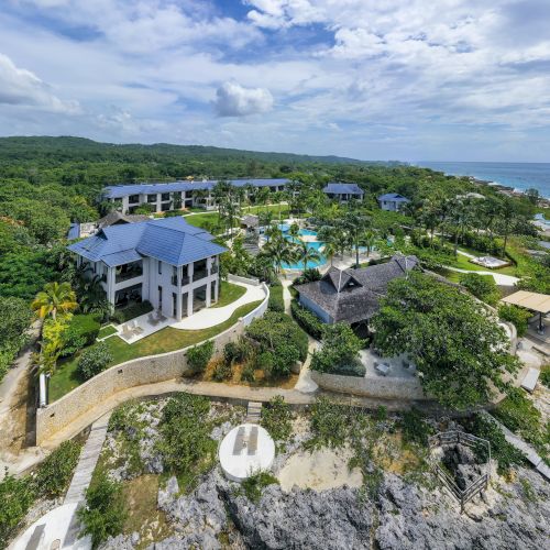 A scenic cliffside overlooking a blue sea with a small white building perched on top, rugged rocks, and green vegetation, under a bright sky.