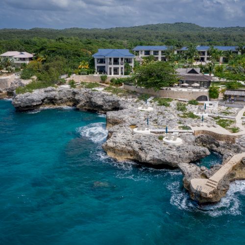A rocky coastline with clear blue water, small coastal buildings, and a bright sunny sky, overlooking a calm cove.