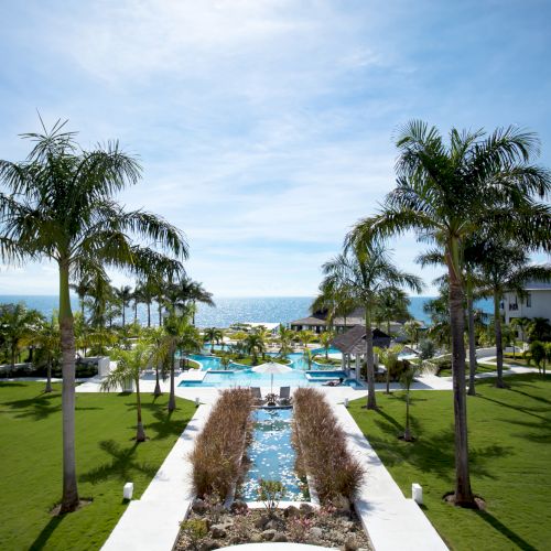 A sunny resort garden with a flagstone path leading to a grand entrance, surrounded by lush green lawn and palm trees.