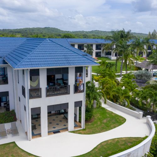 A modern two-story blue-gray house with a balcony, surrounded by a lush tropical lawn and palm trees, under a sunny sky.