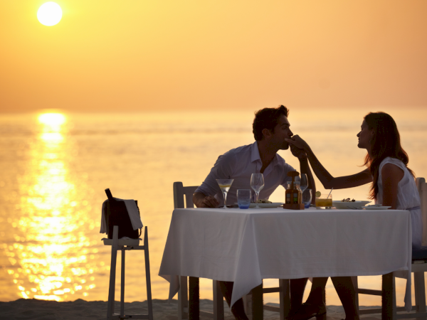 A couple sharing a romantic sunset dinner on the beach, toasting with wine as waves glow in the golden hour.