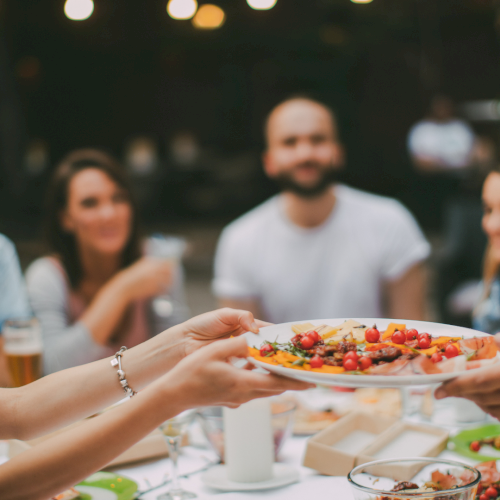 A group of friends sharing a dinner party outdoors, passing a plate of colorful nachos or tapas amid drinks and plates on the table.