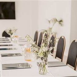A neatly set dining table with white chairs, gold accents, and a bright, elegant centerpiece leading to a sunlit, stylish celebration vibe.