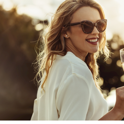 A woman in sunglasses smiles while holding a champagne flute outdoors at sunset, wearing a white blouse.