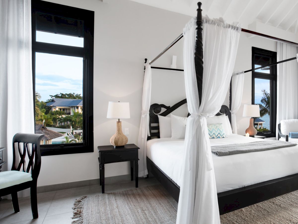 A bright, modern hotel room with a four-poster bed, white linens, black furniture, and large windows letting in natural light.