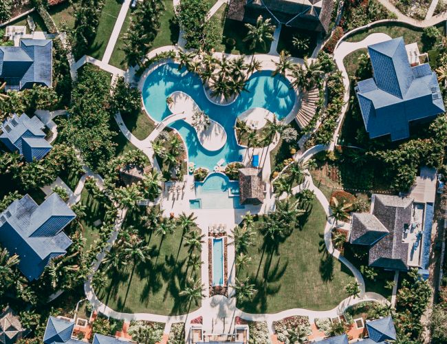 Aerial view of a resort with winding pools, palm trees, and surrounding villas, forming a heart/flower-like layout.