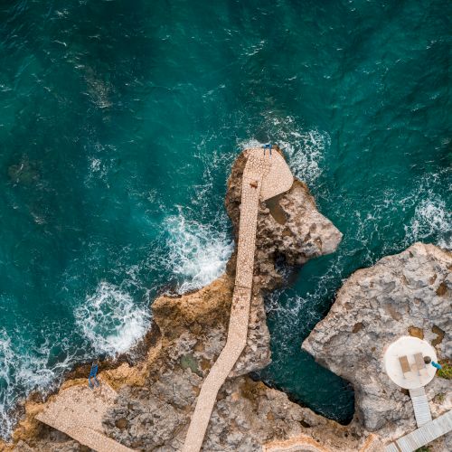 Aerial view of rugged rocky coastline with a concrete jetty extending into turquoise water, small white circular platforms at the edge, waves crashing below.